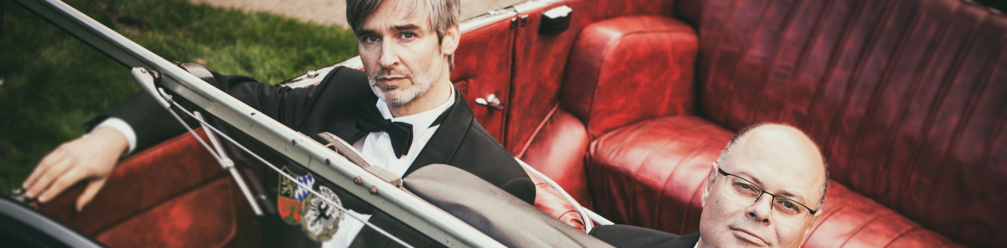 Zwei Musiker des Ballhausorchesters in Frack und Fliege in einem Oldtimer-Cabriolet, &copy; Ballhaus Orchester